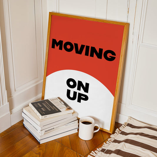 Framed poster with 'MOVING ON UP' text on a stack of books and a cup of coffee in a room.